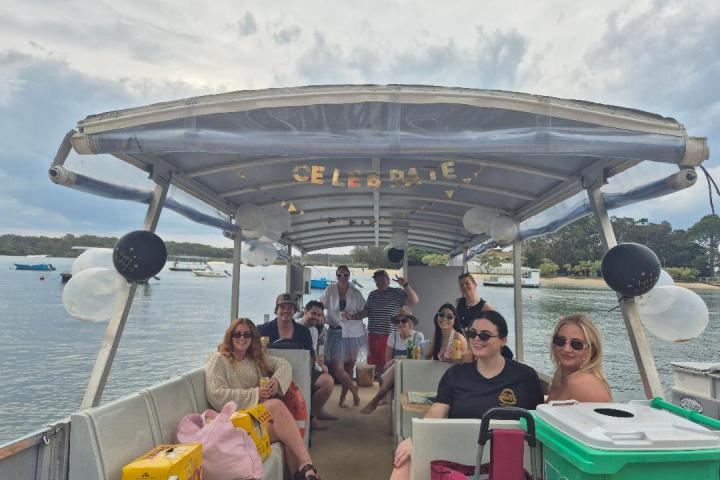 Group of people on a boat with balloons and 'Celebrate' banner, on a scenic river.