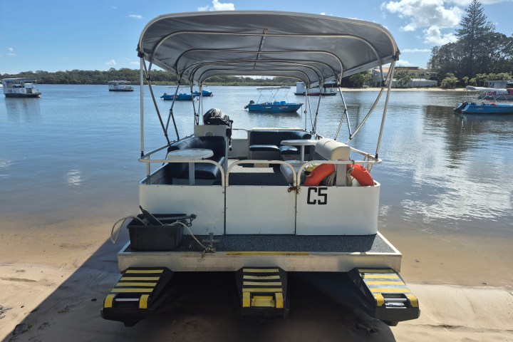 Pontoon boat with canopy docked on sandy shore, calm water and boats in background.