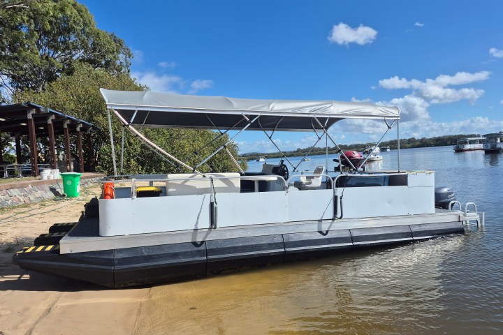 Pontoon boat with canopy docked on sandy shore, sunny day, blue sky.