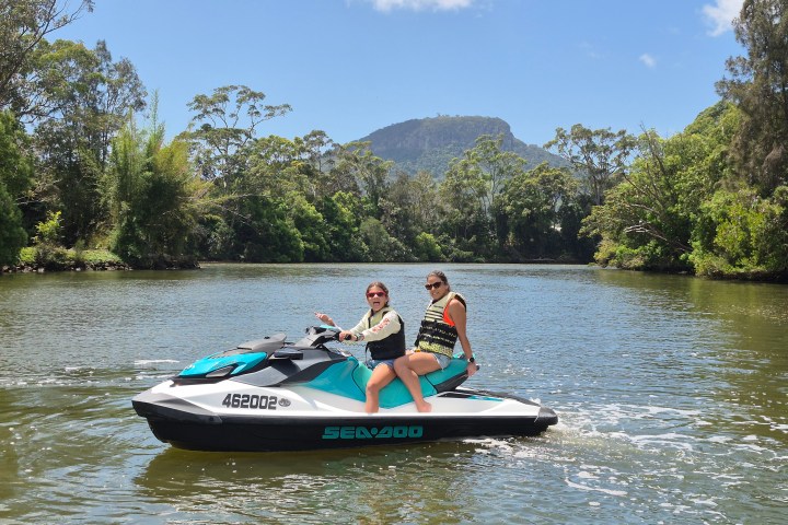 Two people on a jet ski in a river, surrounded by trees and distant hills under a clear blue sky.