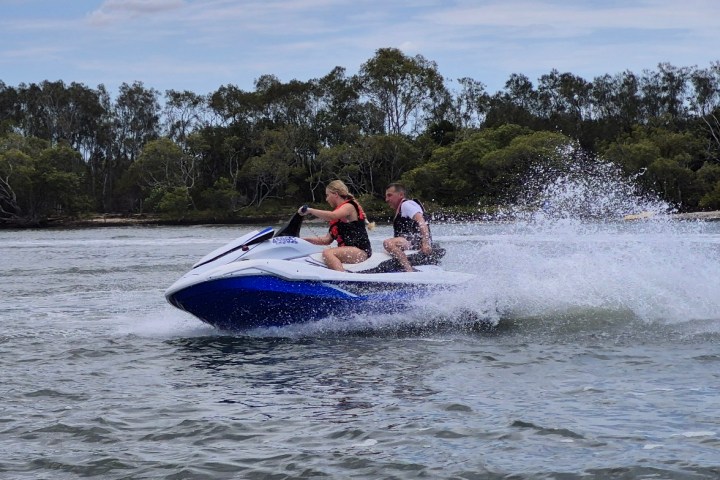 Two people riding a blue jet ski on a lake with trees in the background.