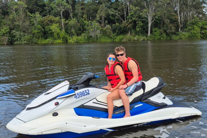 Two people in life vests on a jet ski on a river with trees in background.