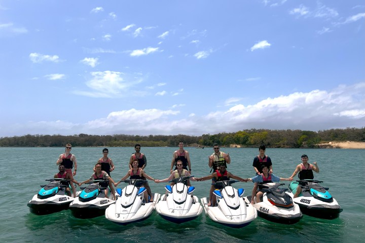 Group of people on jet skis, holding hands on a sunny day with blue sky and trees in the background.