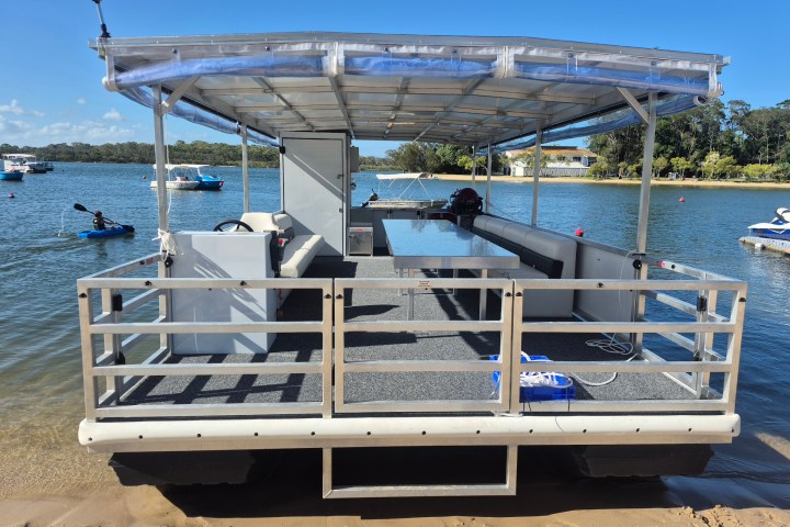 Pontoon boat docked on a sandy shore with a covered deck and railing.