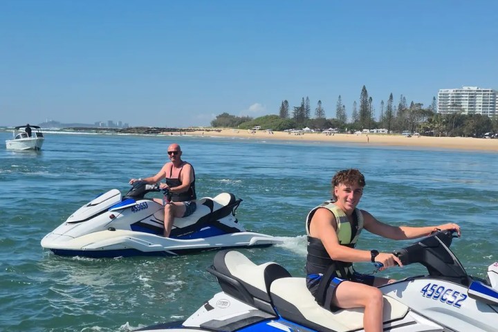 Two people riding jet skis on clear waters near a sandy beach.