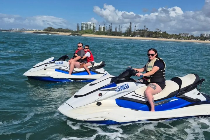 Two people riding jet skis on a sunny day with a beach in the background.